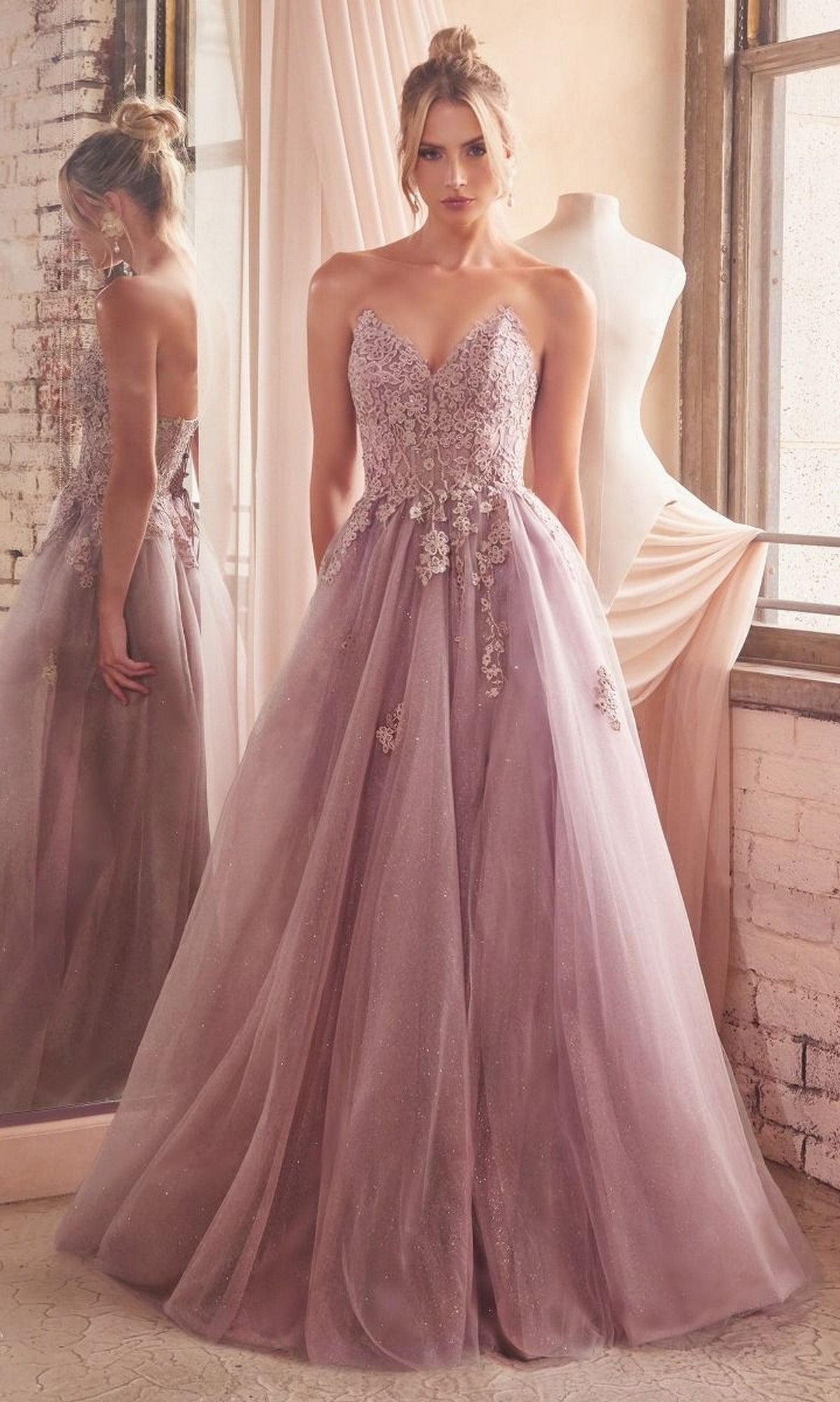 A woman in the Strapless Long Glitter-Tulle Prom Ball Gown C148, featuring an embroidered-lace bodice and flowing tulle skirt, stands by a window and a dress form.